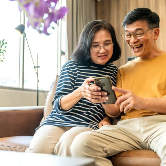 happy senior old asian lover couple holding smartphone looking cellphone screen laughing casual relaxing sit sofa together smiling elder mature grandparents family embracing lifestyle