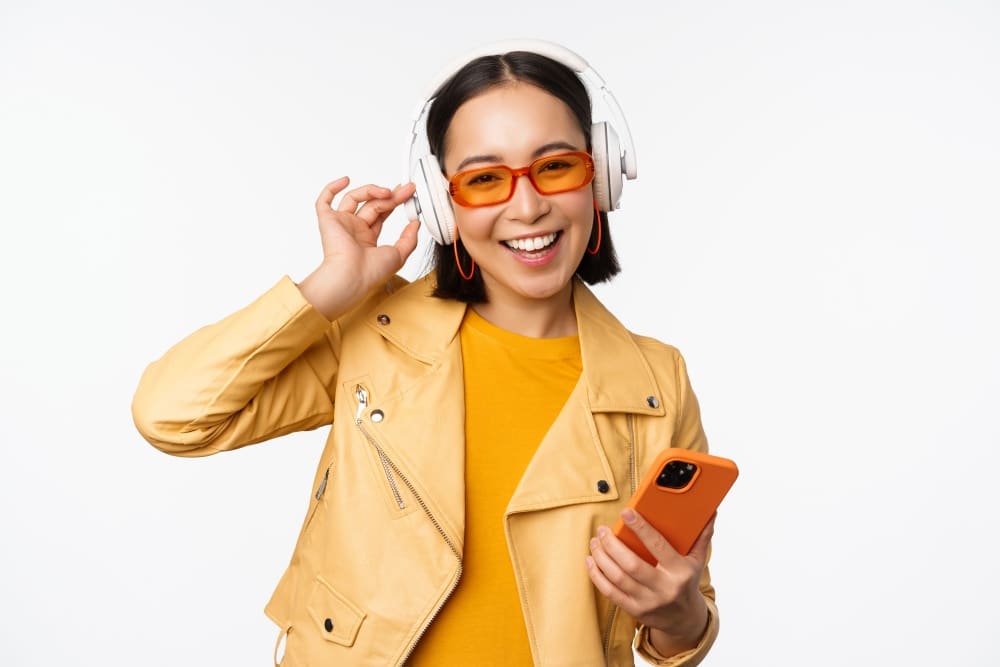 Happy stylish asian woman in sunglasses, listening music in headphones, holding smartphone, singing and dancing, standing over white background