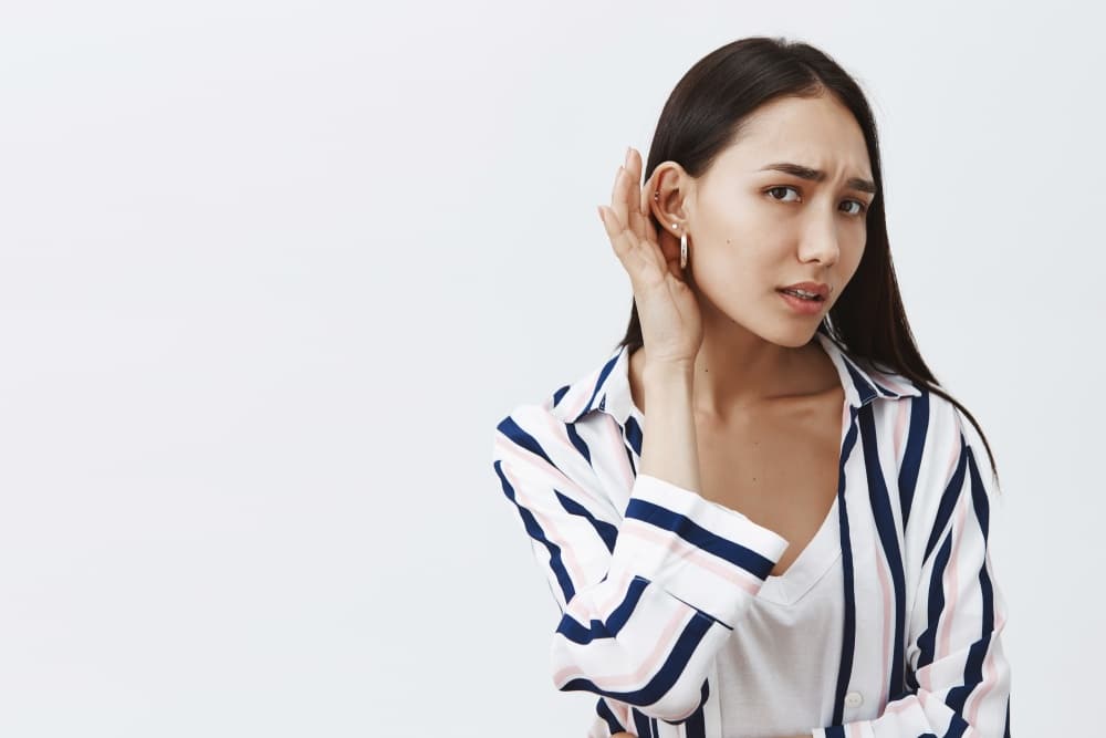 Studio shot of good-looking gloomy girlfriend with long dark hair, leaning towards camera with one ear and gazing at camera, eavesdropping and whispering, hearing rumor, feeling nervous